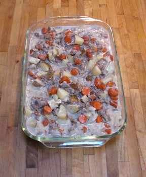 A 9x13 inch glass baking dish filled with ground beef hash with potatoes and carrots, sitting on a maple cutting board counter top.