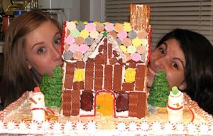 Two girls peeking out from behind a small gingerbread house.