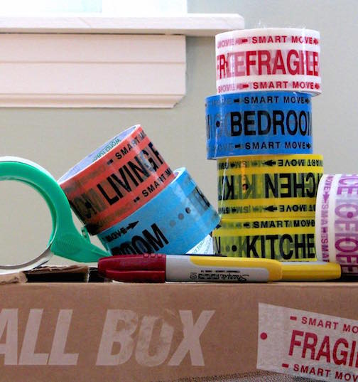 A moving box with rolls of packing tape and markers on it.